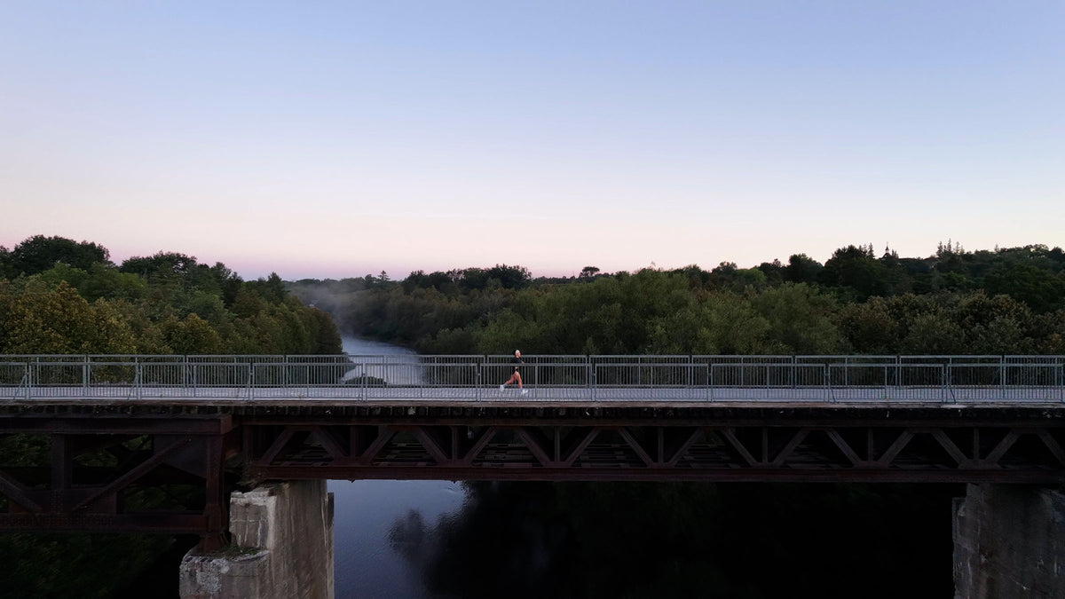 A scenic landscape photograph of a runner running on a bridge pedestrian over a river, surrounded by lush green trees under a soft pastel sky at dawn or dusk.